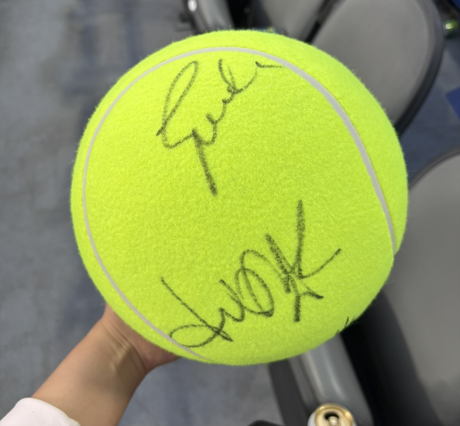 A tennis ball signed by Iga Świątek, Shuai Zhang, and Kateřina Siniaková, held beside the stadium seats.